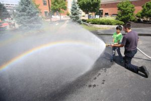 Water coming from a fire hose held by Forest and a firefighter, makes a rainbow