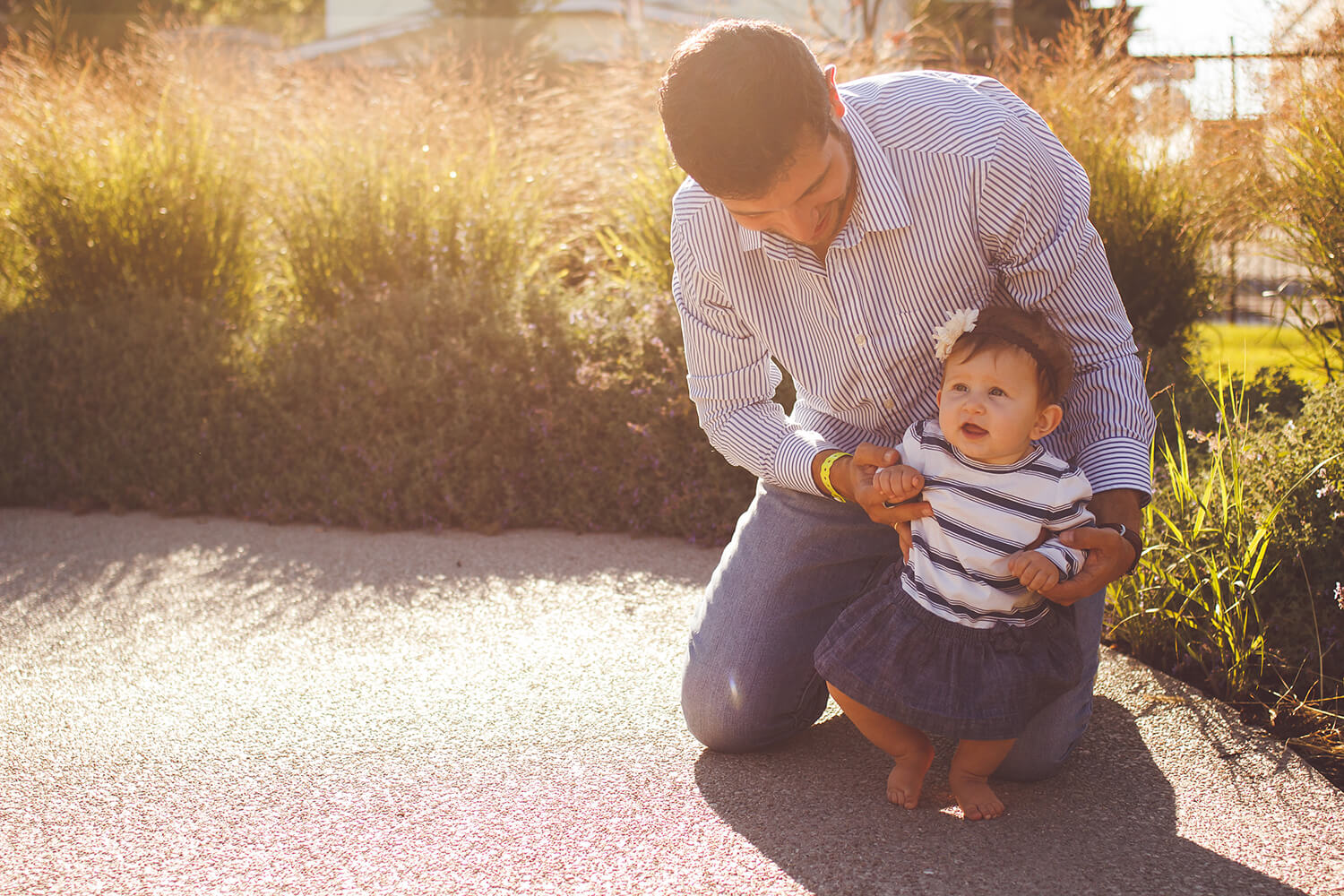 Baby girl is held up by her dad who is kneeling behind her as the sun sets golden light behind them.