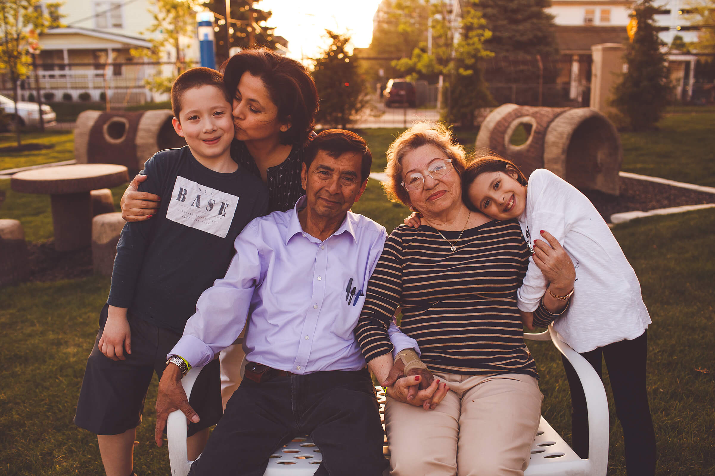 A grandmother and grandfather sit on a bench holding hands. A young girl hugs onto her grandmothers arm. A young boy and the mother stand behind the bench, as the mother kisses her sons cheek. It's around the time of sunset. The whole family is smiling.