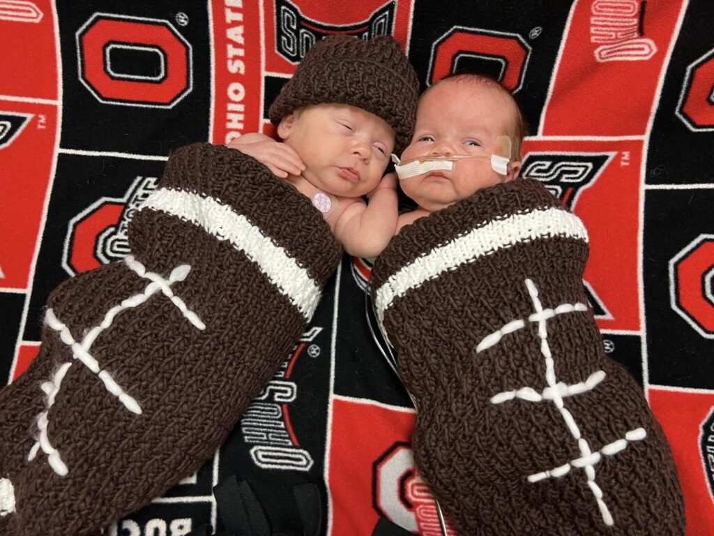 Premature twin babies are swaddled in knitted footballs while laying on an Ohio State University blanket.