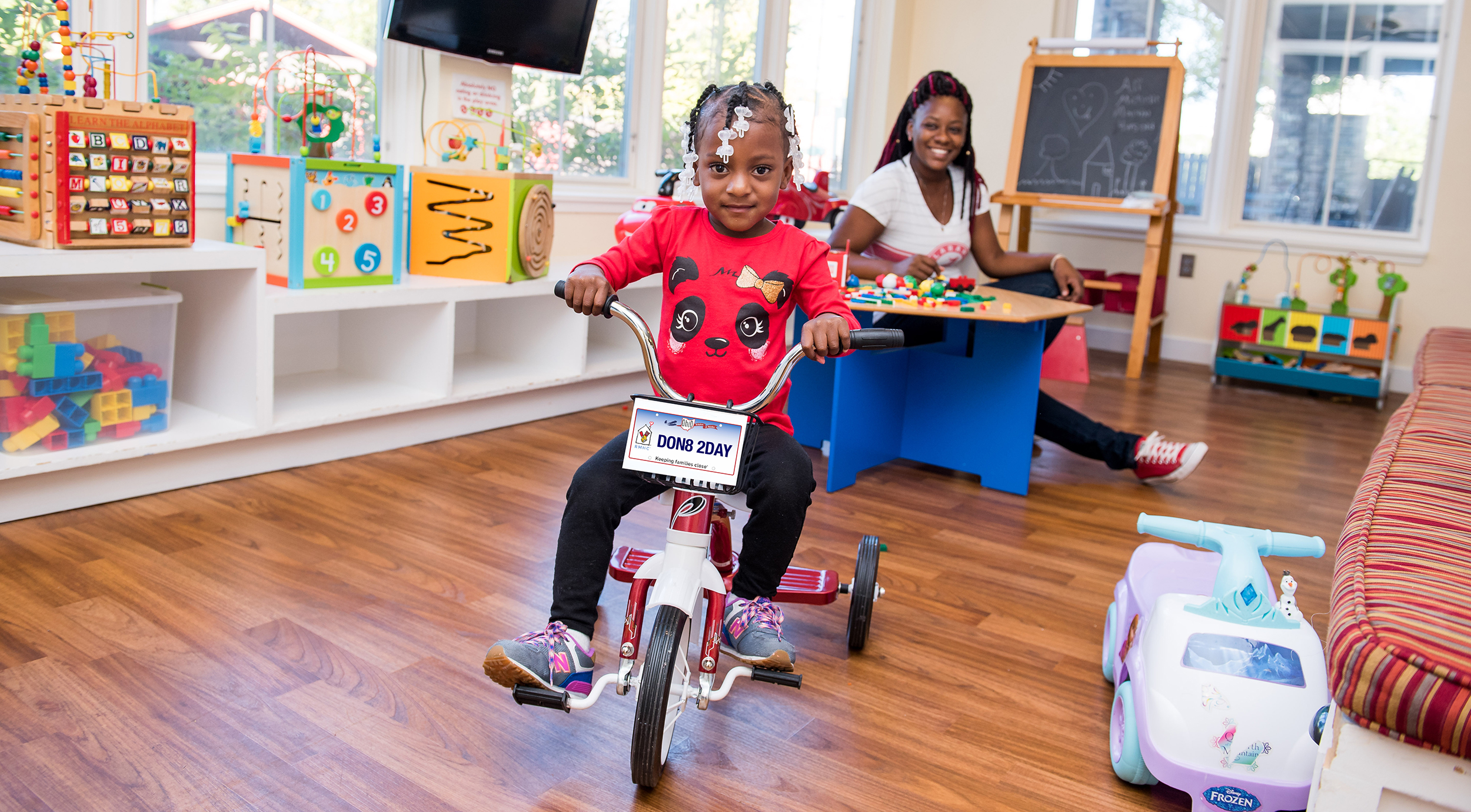 A child riding a tricycle in a playroom full of toys, smiling while her mom smiles and watches her in the background.