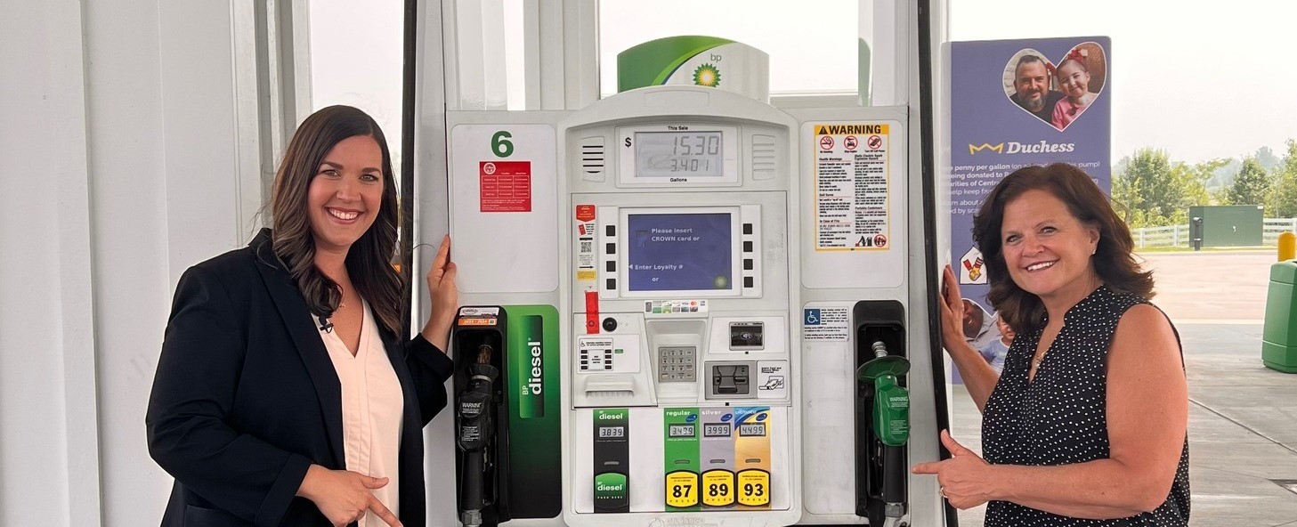 Two people stand on either side of a gas pump pointing to it.